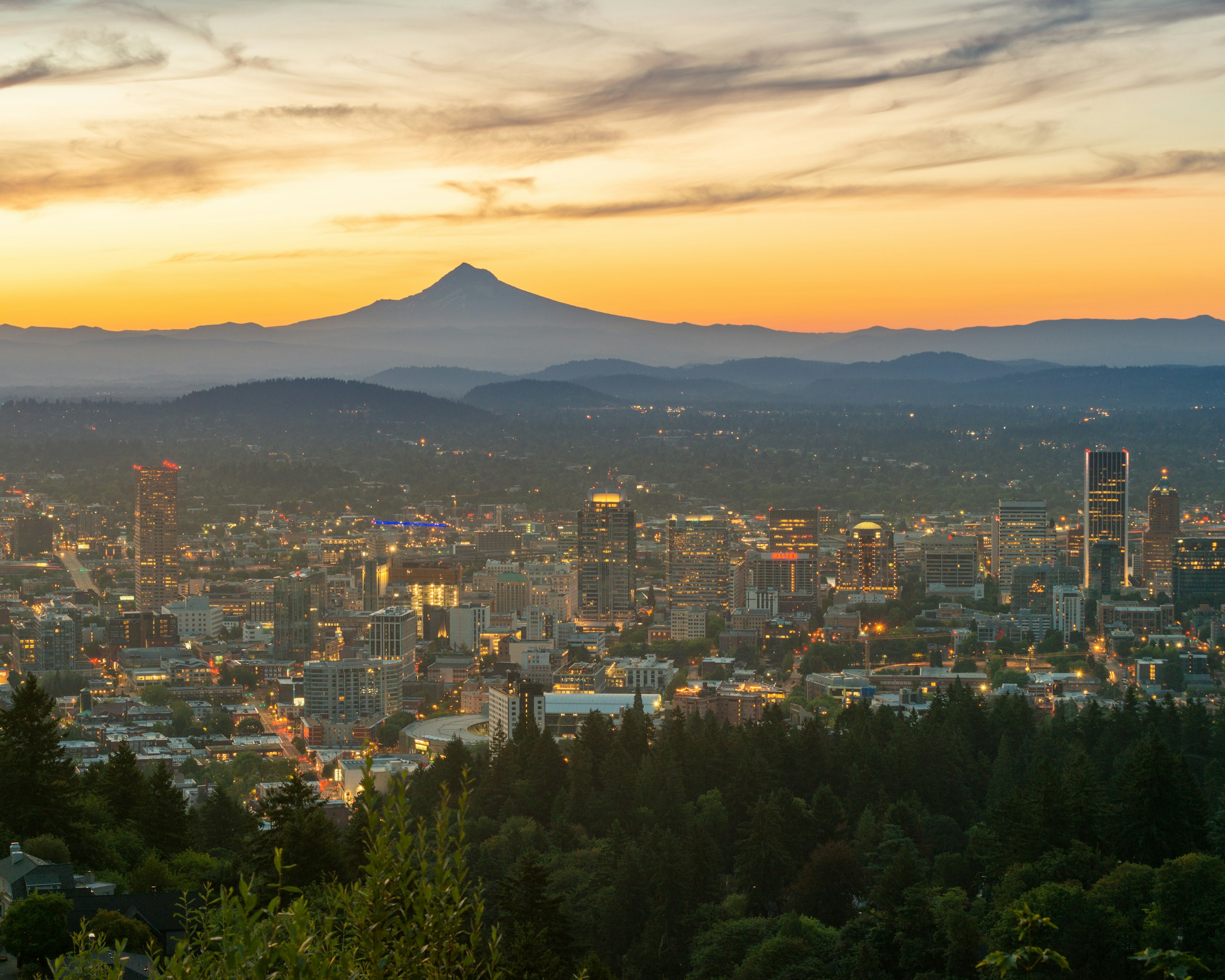 Portland rooftops and forested hills with Mount Hood beyond, in warm evening light.